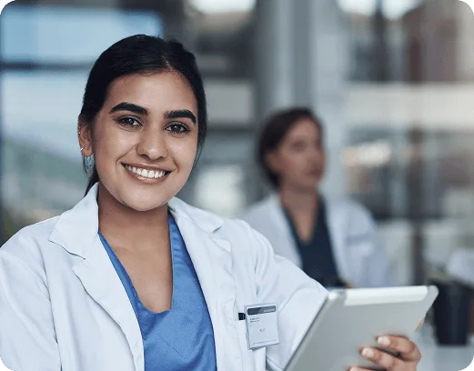 ambulance image 58784 webp Smiling woman in white coat over blue scrubs holding a tablet, colleague blurred behind