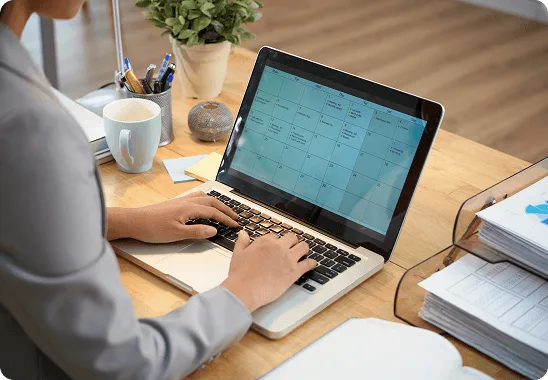 Person typing on a laptop showing a calendar at a desk with coffee mug and paper trays