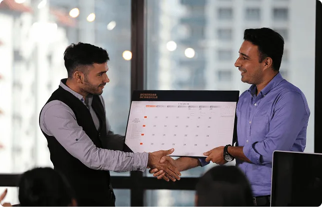 Two men in an office shaking hands while holding a printed business schedule calendar between them