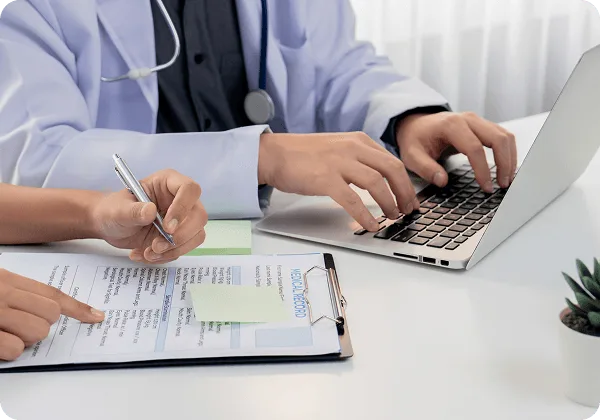 Clinician in a white coat typing on a laptop while another hand points at a record form
