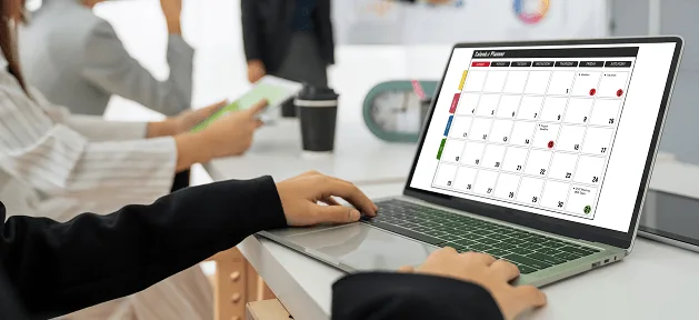 Person typing on a laptop displaying a monthly calendar planner, with colleagues and coffee cup nearby