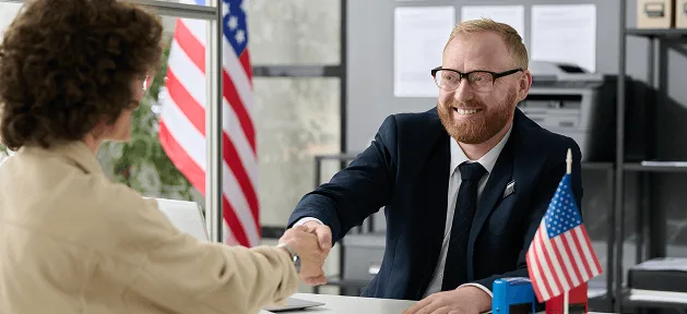 Smiling bearded man with glasses in a suit shaking hands with a person across a desk