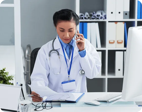 Woman in a white coat and stethoscope on a phone call reviewing papers at a desk