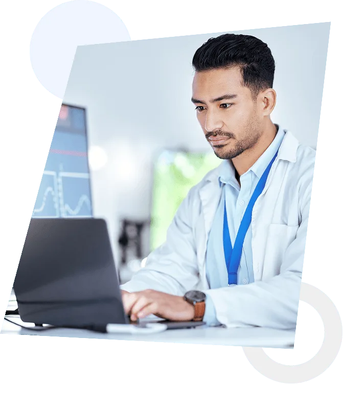 Man in white coat and blue lanyard typing on a laptop in a bright office