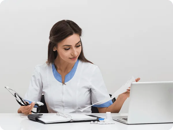 Female healthcare worker at a desk holding a stethoscope and paper beside a laptop