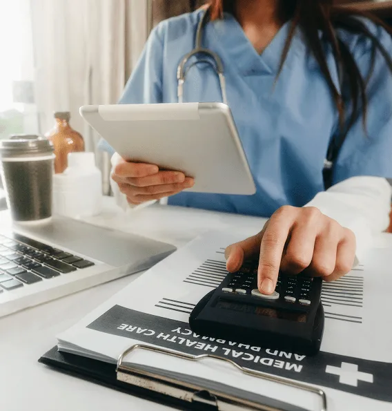Healthcare worker in scrubs holding a tablet and pressing a calculator on a medical and pharmacy clipboard