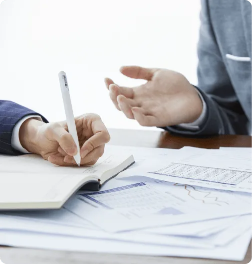 Close-up of a hand writing in a notebook beside printed charts and papers while another person gestures