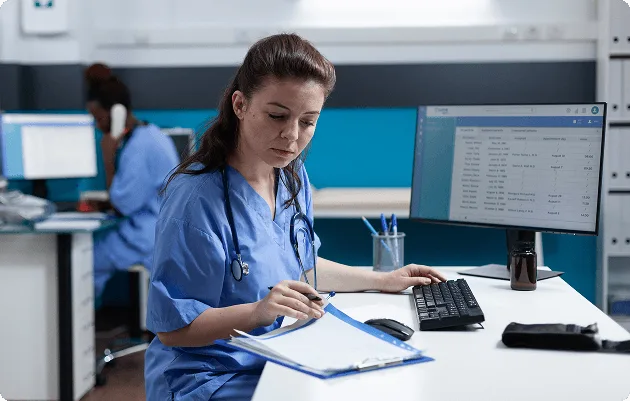 Nurse in blue scrubs with stethoscope writing on a clipboard at a desk with a monitor