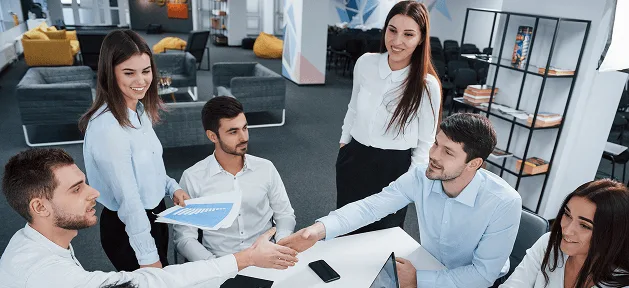 Group of colleagues in a bright office smiling and shaking hands around a table with charts and a laptop