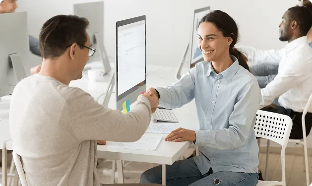 Smiling woman shaking hands with a man at a desk with computer monitors and coworkers in the background