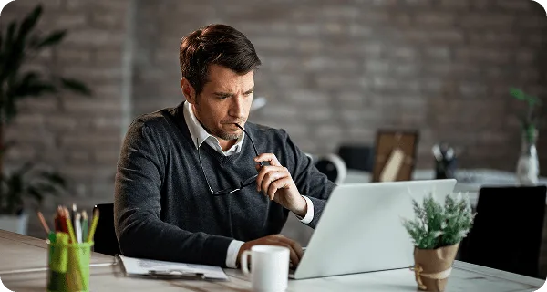 Man in sweater holding glasses to his mouth while looking at a laptop at a desk