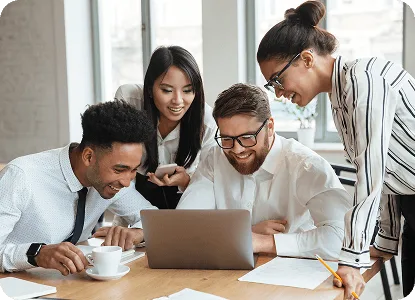 Four colleagues in office clothes smiling and gathered around a laptop on a table