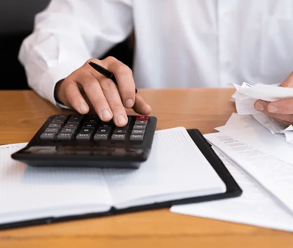 Hand using a calculator on a desk with a notebook, pen and scattered paperwork