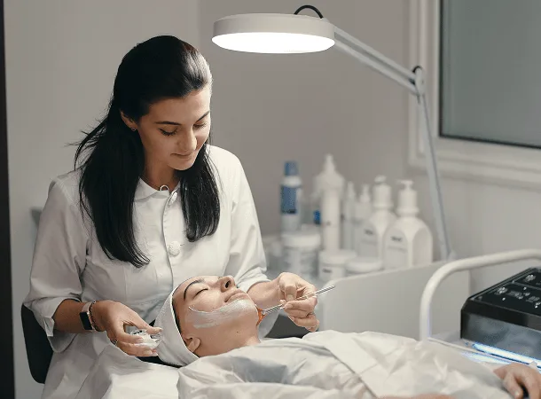 dermatology image 59933 webp Skincare specialist applying a facial mask to a reclining client under a bright lamp