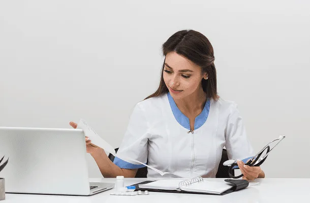 Woman in white medical tunic reading a document at a desk with laptop and stethoscope