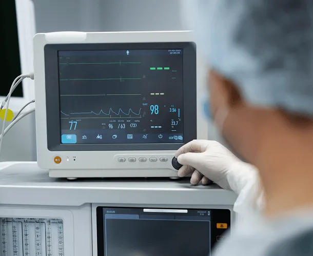 Gloved hand adjusting a dial on a patient monitor showing vital sign waveforms