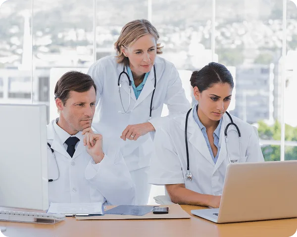 Three clinicians in white coats with stethoscopes reviewing a laptop and monitor in a bright office