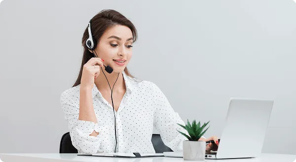 Smiling woman wearing a headset working at a laptop with a small potted plant on a white desk
