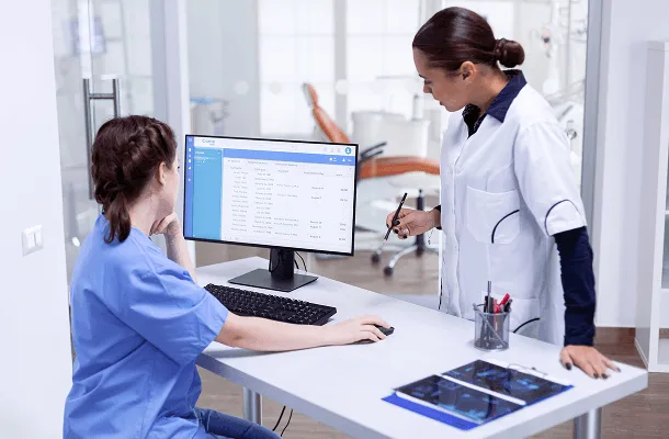 Two women in scrubs and a white coat look at a desktop monitor showing a data table in a clinic office