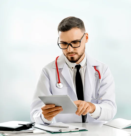 Doctor in white coat and glasses with red stethoscope reading a tablet at a desk