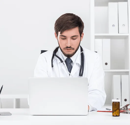 Man in white coat with stethoscope working at a laptop in an office with binders