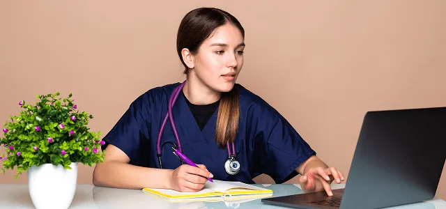 Woman in navy scrubs with stethoscope writing in a notebook while using a laptop