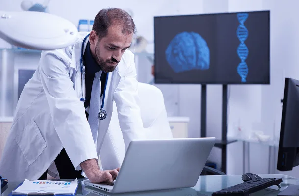 gastroenterology image 59884 webp Bearded man in white coat and stethoscope leans over a laptop, with brain and DNA display behind