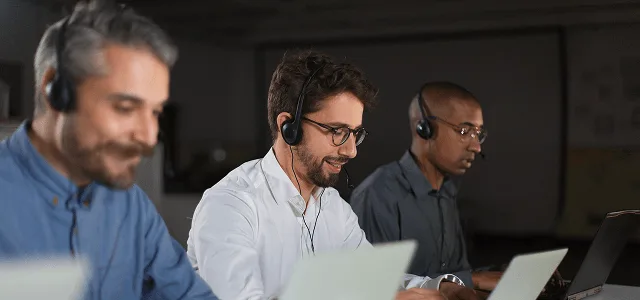 gastroenterology image 59888 webp Three men wearing headsets smile while working on laptops in a dim office