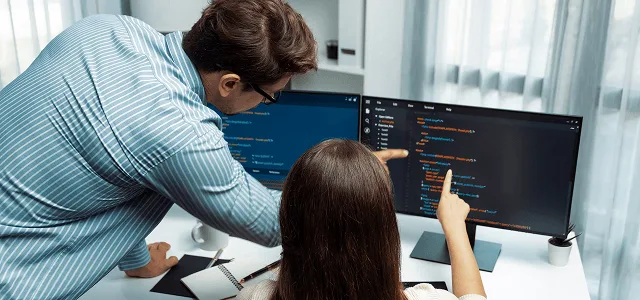 Two coworkers at a desk pointing at dark computer screens displaying lines of code