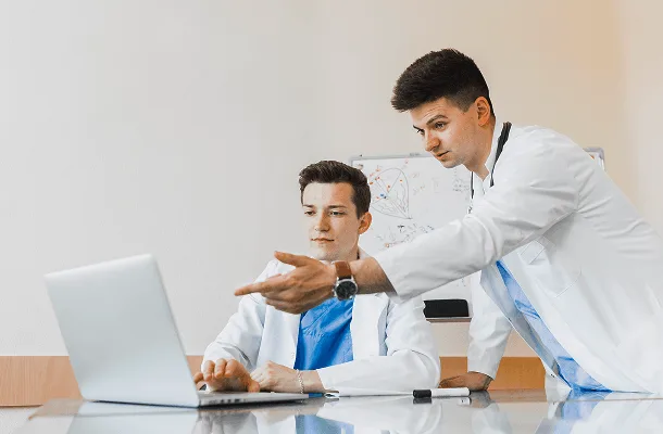 Two doctors in white coats looking at a laptop, one standing and pointing at the screen