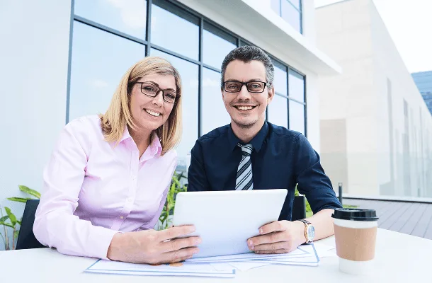 Smiling woman and man with glasses sitting outdoors holding a tablet with papers on the table