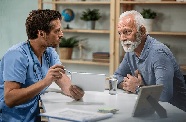 geriatrics image 59867 webp Clinician in scrubs with a stethoscope speaks with an older bearded man across a table