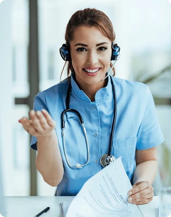 Smiling woman in blue scrubs wearing a headset and stethoscope, holding papers at a desk