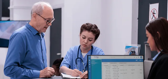 Nurse in scrubs writing notes while an older man stands beside her at a reception desk