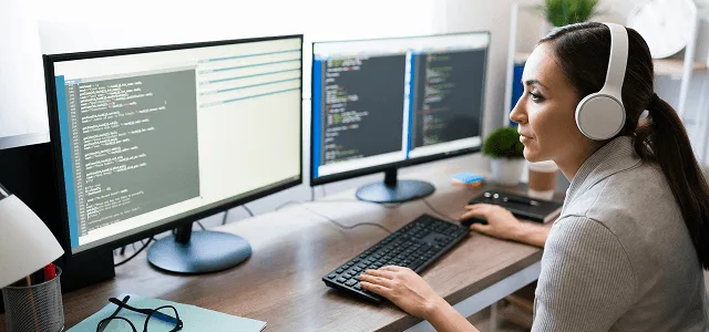 Woman wearing headphones working at a desk with two monitors showing code