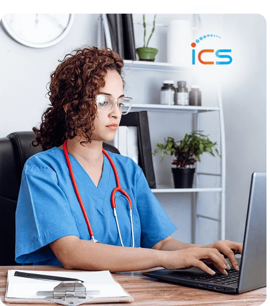 Woman in blue scrubs with red stethoscope typing on a laptop at a desk