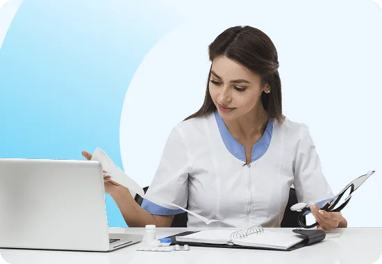 Woman in a white and blue medical uniform reviewing papers at a desk with a laptop