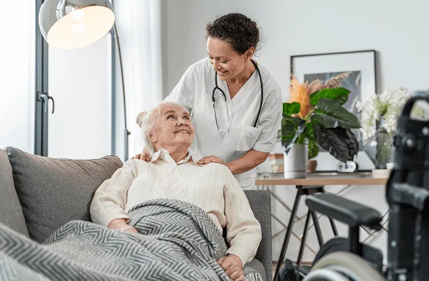 Smiling nurse with a stethoscope leaning over an elderly woman resting on a sofa at home