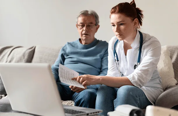 Doctor and older man on a sofa looking at a laptop while she points to a paper
