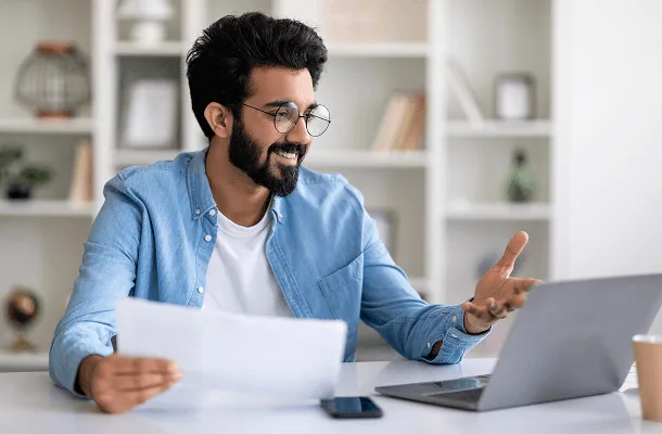 Smiling bearded man in glasses holding a paper and gesturing toward his laptop at a desk