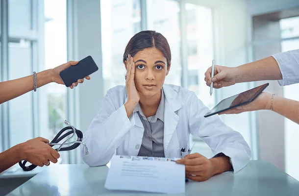 Stressed doctor with hand on head while several hands hold out a phone, tablet, pen and stethoscope