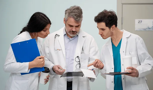 Three medical professionals in white coats reviewing papers and a clipboard together