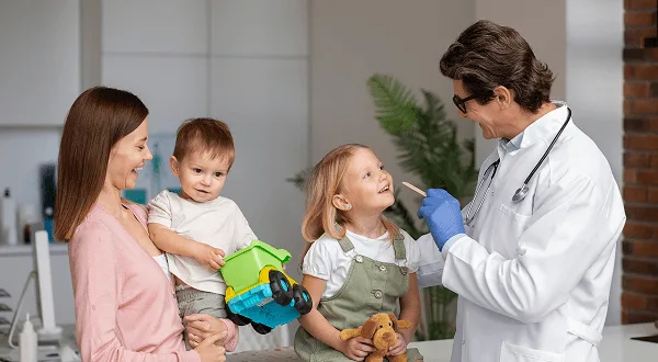 Doctor in a white coat with gloves examining a young girl while a mother holds a toddler