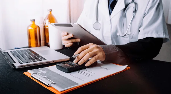 Healthcare worker using a calculator at a desk with a tablet, laptop and clipboard
