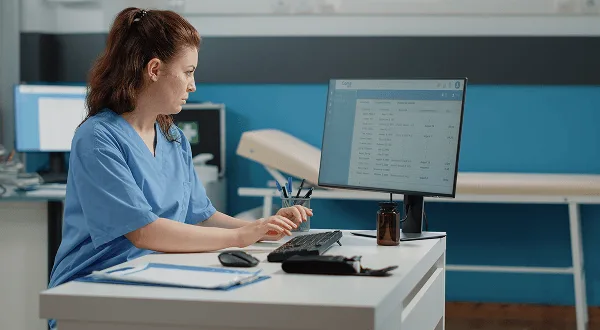 Nurse in blue scrubs typing at a desktop computer showing a patient list