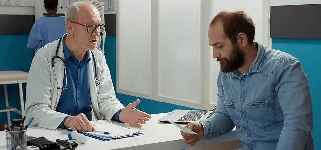 Doctor in a white coat talking with a seated patient at a desk in a clinic