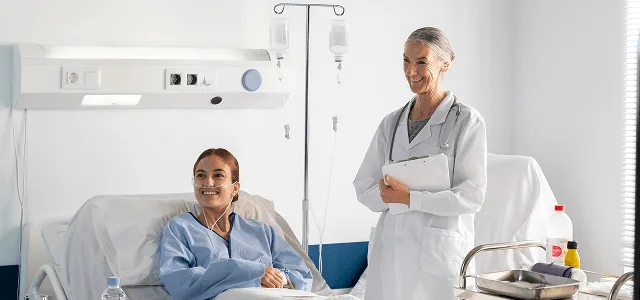 Smiling patient in a hospital bed with a smiling doctor holding a clipboard beside an IV stand