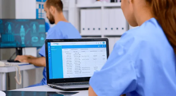 Two staff members in blue scrubs viewing a patient schedule on a laptop