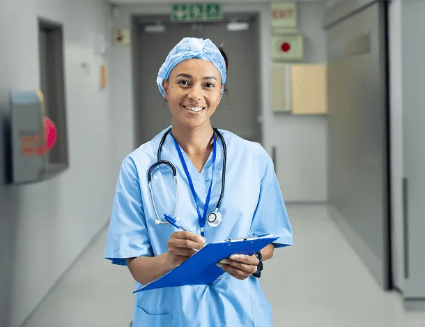 Smiling woman in light blue scrubs and surgical cap holds a clipboard and pen in a hospital corridor