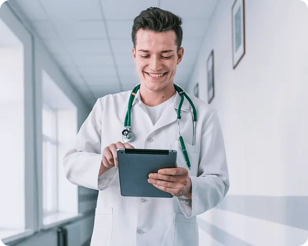 Smiling man in a white coat with a stethoscope looking at a tablet in a hospital corridor
